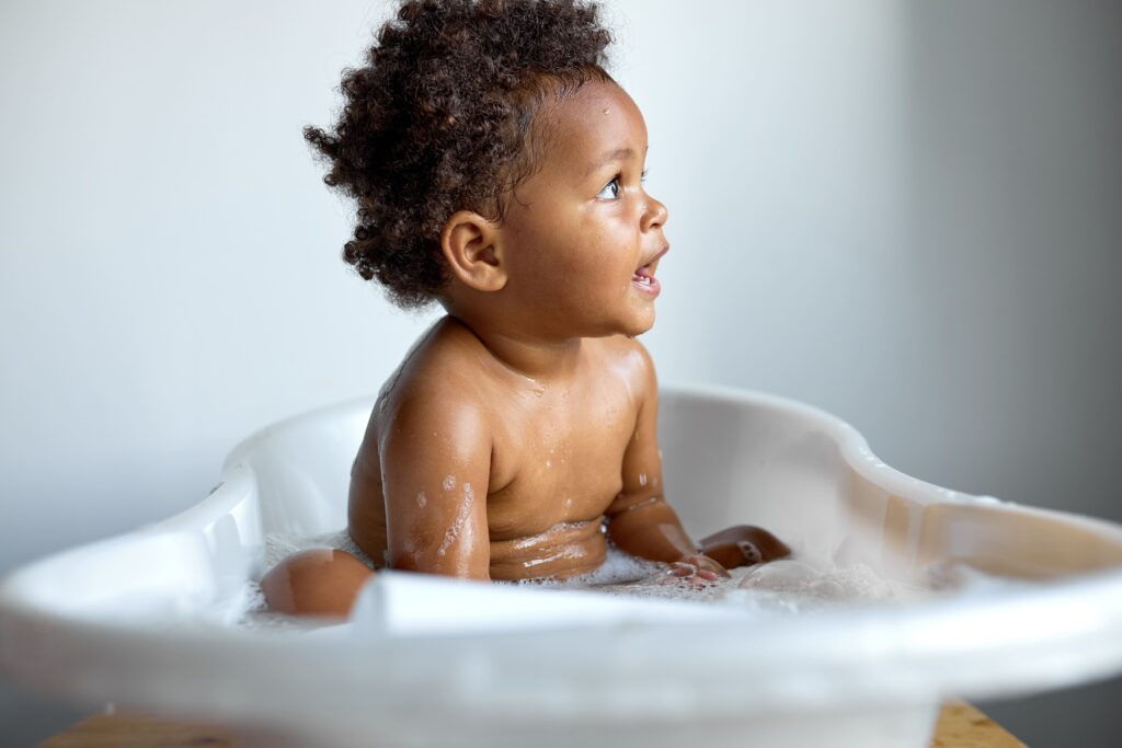 Small cute black child sitting at bath, washing, playing with soap and foam, indoors, copy space, white background, happy childhood, banner. adorable kid enjoying hygiene skincare procedures